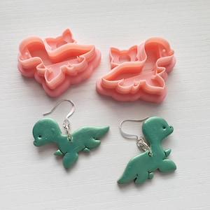 May include: Two pairs of green dinosaur earrings with silver hooks. The earrings are made of polymer clay and are shaped like dinosaurs. There are also two pink dinosaur cookie cutters in the image.