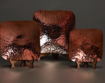 May include: Three hammered copper serving trays with wooden stands. The trays are square and have a hammered texture. The stands are made of wood and have three legs.