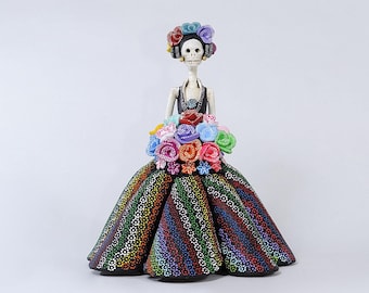 May include: A Day of the Dead figurine of a skeleton woman wearing a black dress with colorful polka dots and a floral crown. She is holding a bouquet of colorful flowers.