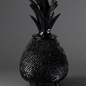 Puede incluir: Un jarrón de cerámica negro con forma de piña con una superficie texturizada y tres patas. El jarrón tiene una parte superior de piña detallada con hojas.