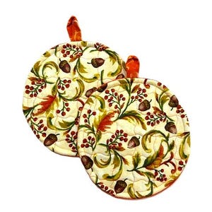 May include: Two round quilted pot holders featuring a cream-colored background with a repeating autumn-themed pattern of brown acorns, green leaves, and clusters of small red berries. Each pot holder includes a small orange fabric hanging loop.