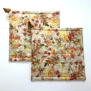 May include: Two square quilted pot holders featuring a repeating autumn pattern of orange, yellow, and red maple and oak leaves, brown acorns, and small red berries on a light beige background, each with a small brown fabric hanging loop.