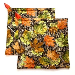 May include: Two square fabric pot holders featuring a realistic forest floor pattern with brown tree bark, green moss, and fallen autumn leaves in shades of orange, yellow, and brown. Each pot holder has a small, bright orange fabric hanging loop.