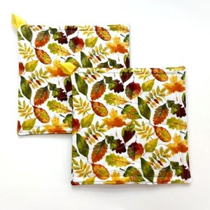 May include: Two square quilted pot holders featuring a white background with a repeating pattern of autumn leaves in shades of orange, yellow, brown, and green. Each pot holder includes a small yellow fabric loop attached to one corner.