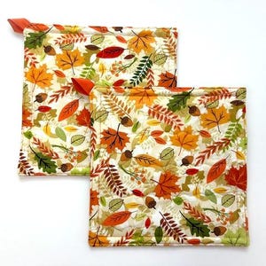 May include: Two square quilted pot holders featuring a cream background with a colorful autumn pattern of orange, red, yellow, and green leaves, acorns, and berries. Each pot holder has a small orange fabric loop attached to one corner for hanging.