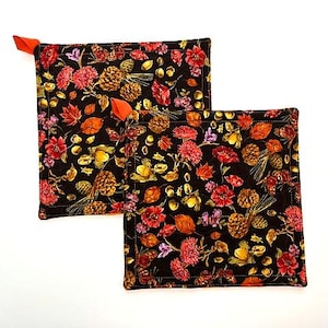 May include: Two square quilted pot holders featuring a black background with a vibrant autumn-themed print of red flowers, orange leaves, brown pinecones, and acorns. Each pot holder includes a small, bright orange fabric hanging loop at the corner.