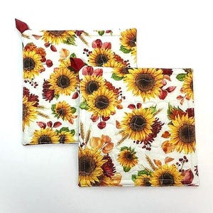 May include: Two square quilted pot holders featuring a white background with a repeating pattern of yellow sunflowers, brown centers, green leaves, wheat stalks, and small red berries. Each pot holder has a small red fabric loop at the corner.