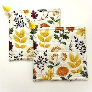 May include: Two square quilted pot holders featuring a white background with a repeating pattern of yellow leaves, orange chrysanthemums, dark green foliage, and small dark berries. Each pot holder has a small yellow fabric loop at the corner.