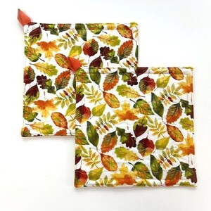 May include: Two square quilted pot holders featuring a white background with a repeating pattern of autumn leaves in shades of orange, yellow, brown, and green. Each pot holder has a small orange fabric loop attached to one corner for hanging.