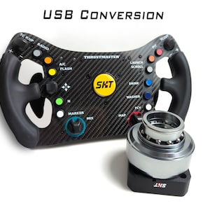 May include: A black carbon fiber Thrustmaster racing steering wheel with a USB CONVERSION label, featuring numerous colorful buttons, a central yellow SKT logo, and a separate silver metal quick-release adapter with a black base and SKT logo.