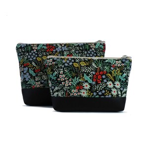 May include: Two black zippered pouches with a floral print. The larger pouch is behind the smaller pouch. Both pouches have a black leather bottom.