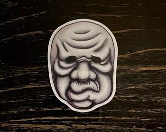 May include: A sticker featuring a detailed illustration of a Noh mask. The mask is primarily gray with darker shading to define the features, including the eyes, nose, and mouth. The mask has a slightly furrowed brow and a stern expression. The sticker is on a dark wooden surface.
