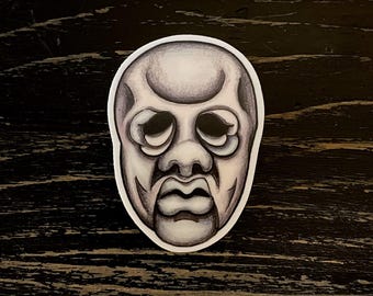 May include: A grayscale sticker featuring a stylized, theatrical mask with a prominent brow, large eyes, and a defined mouth. The mask has a slightly distressed appearance, with shading that gives it depth and texture. The sticker is on a dark wood-grain surface.
