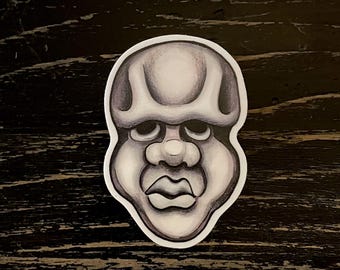 May include: A grayscale sticker featuring a stylized face with prominent features. The design includes a large forehead, heavy brow, a rounded nose, and full lips. The sticker has a white border and is set against a dark wood grain background.