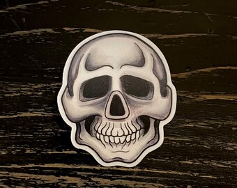 May include: A grayscale skull sticker with detailed shading, featuring a prominent nose cavity, eye sockets, and a full set of teeth. The sticker has a white border and is set against a dark, textured background.