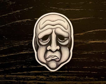 May include: A sticker featuring a grayscale illustration of a sad face. The face has a rounded shape with heavy, furrowed brows, downturned eyes, and a small, downturned mouth. The sticker is outlined in white.