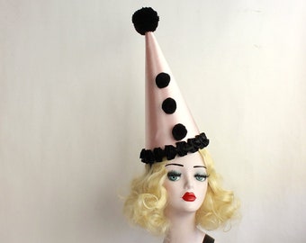 Puede incluir: Un sombrero de cono rosa con un pompón negro en la parte superior y tres pompones negros en la parte delantera. El sombrero tiene un volante negro alrededor de la base.