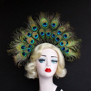 May include: A mannequin head with blonde hair wearing a peacock feather headdress. The headdress is made of green and blue feathers with iridescent eyes.