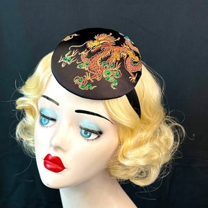 May include: A black satin hat with a round, flat top featuring an embroidered gold dragon with green accents.