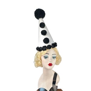 May include: A white cone-shaped hat with black pom-poms and a black trim. The hat is worn by a mannequin with blonde hair.