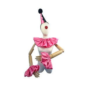 Puede incluir: Un conjunto de disfraz de payaso sobre un maniquí. El conjunto incluye un cuello con volantes de satén rosa, puños y un sombrero de cono a juego con lunares negros y un pompón negro. El maniquí tiene una nariz roja.