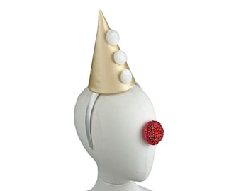 May include: A gold and white clown hat with three white pom-poms on a white mannequin head with a red rhinestone nose.