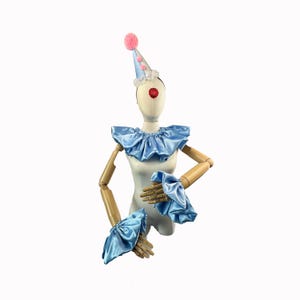 May include: A clown costume set on a mannequin. The set includes a light blue satin ruffled collar, wrist cuffs, and a cone hat with pink pom-pom and white lace trim. A red nose is also included.