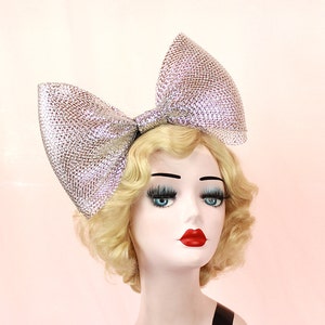May include: A large silver sparkly bow headband. The bow is made of a glittery material and is tied in a large, dramatic knot. The headband is worn on a mannequin with blonde hair.