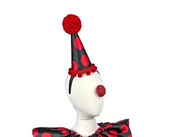 May include: A black and red clown hat with red pom-poms and a red nose. The hat is on a white mannequin head with a red and black polka dot collar.