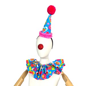 May include: A blue and pink clown hat with a pink pom-pom on top and a ruffle around the base. The hat has a pattern of colorful hearts. A red clown nose is attached to a white mannequin head. The mannequin is wearing a blue and pink ruffled collar with a pattern of colorful hearts.