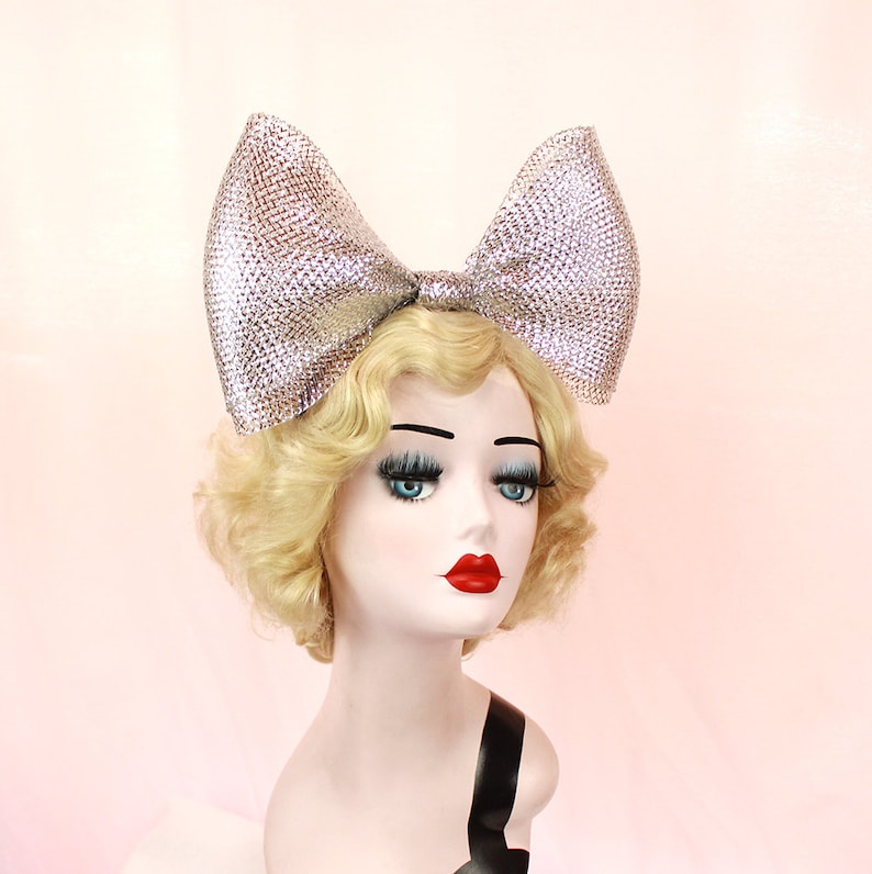 May include: A large silver sparkly bow headband. The bow is made of a shiny, textured material and is tied in a large, dramatic knot. The headband is worn on a mannequin with blonde hair.