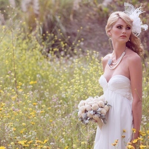 May include: A bride in a white strapless wedding dress with a white flower and feather headpiece holds a bouquet of white flowers in a field of yellow wildflowers.