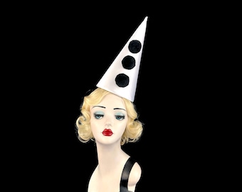 May include: A white conical party hat featuring three vertically aligned black pom-poms, displayed on a mannequin head with blonde hair, red lips, and blue eyeshadow, set against a solid black background.