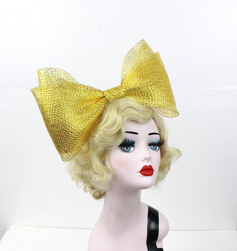 May include: A large gold mesh bow hair accessory. The bow is made of a sparkly gold mesh fabric and is tied in a large, dramatic bow.