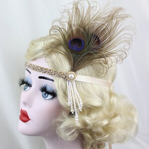 May include: A gold headband with a row of rhinestones and a pearl accent. A single peacock feather with blue and green eyes is attached to the headband. A strand of pearls hangs from the pearl accent.