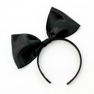 May include: A black satin bow headband. The headband is made of a thin black elastic band with a large black satin bow attached to the top.