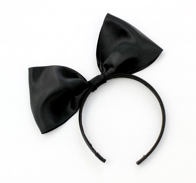 May include: A black satin bow headband. The headband is made of a thin black elastic band with a large black satin bow attached to the top.