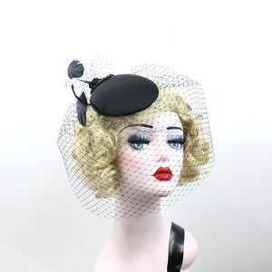 May include: A black pillbox hat with a black feather and a black veil. The hat is sitting on a mannequin head with blonde hair.