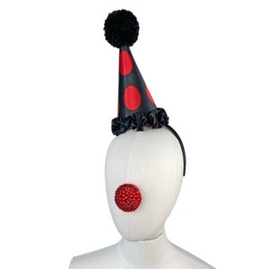 May include: A black and red polka dot party hat with a black pom pom on top. The hat is on a white mannequin head with a red jeweled clown nose.
