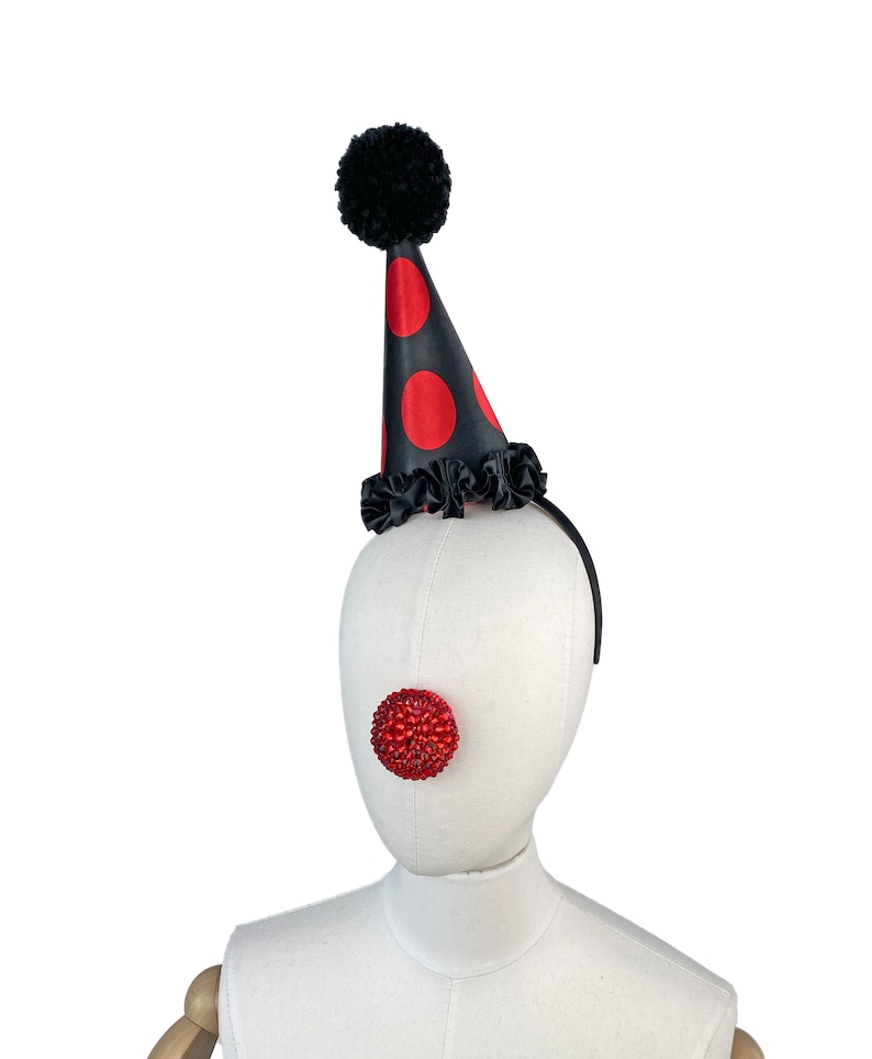 May include: A black and red polka dot party hat with a black pom pom on top. The hat is on a white mannequin head with a red jeweled clown nose.