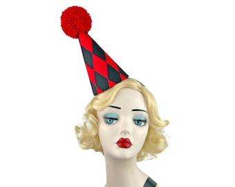 May include: A red, black, and white harlequin-patterned party hat with a red pom-pom on top. The hat is worn by a mannequin head with blonde hair, blue eyes, and red lipstick. The hat is a festive accessory.