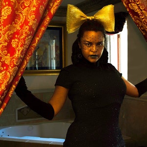 May include: A woman wearing a black dress with a gold bow headband and a black lace veil. She is standing in front of a red and gold patterned curtain.