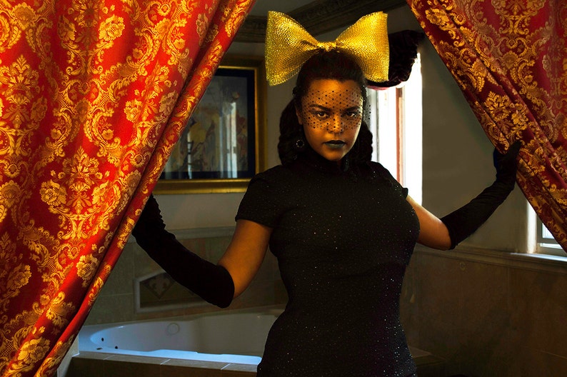 May include: A woman wearing a black dress with a gold bow headband and a black lace veil. She is standing in front of a red and gold patterned curtain.