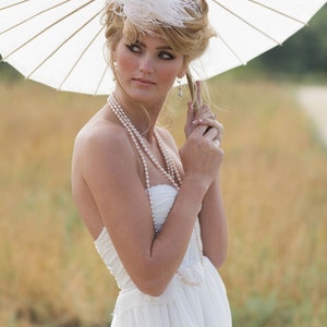 May include: A woman wearing a white strapless dress with a pearl necklace and a white feathered hat. She is holding a white parasol.