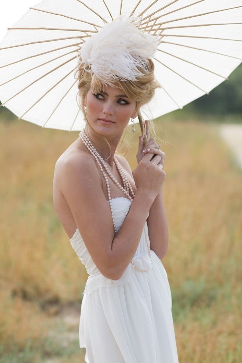 May include: A woman wearing a white strapless dress with a pearl necklace and a white feathered hat. She is holding a white parasol.