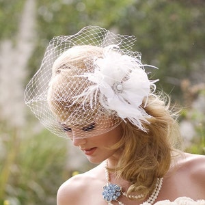 May include: A white bridal veil with a delicate floral design and a single feather accent. The veil is worn by a woman with blonde hair and a pearl necklace.