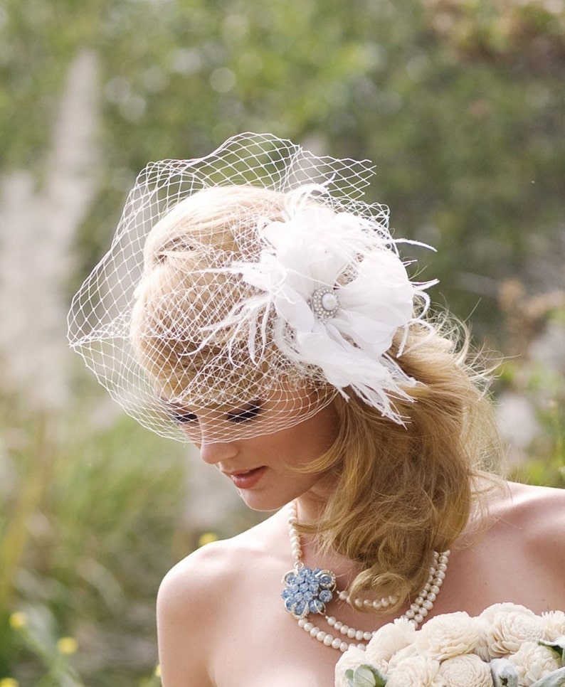May include: A white bridal veil with a delicate floral design and a single feather accent. The veil is worn by a woman with blonde hair and a pearl necklace.