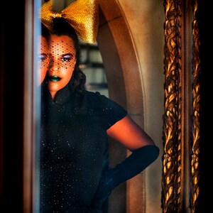 May include: A woman wearing a black dress with a gold bow and a black veil over her eyes. She is standing in front of a mirror with a gold frame.