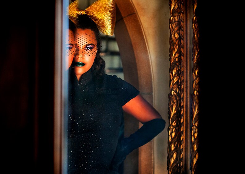 May include: A woman wearing a black dress with a gold bow and a black veil over her eyes. She is standing in front of a mirror with a gold frame.