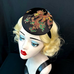 May include: A black satin hat featuring a gold and red embroidered dragon design. The dragon has green accents and is surrounded by gold and green embellishments.